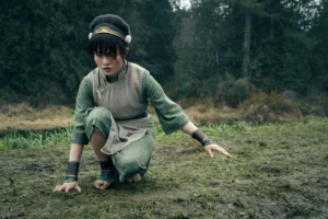 "A young girl with black hair wearing green clothes crouches down in the woods with bare feet on grass and right hand touching the ground. "