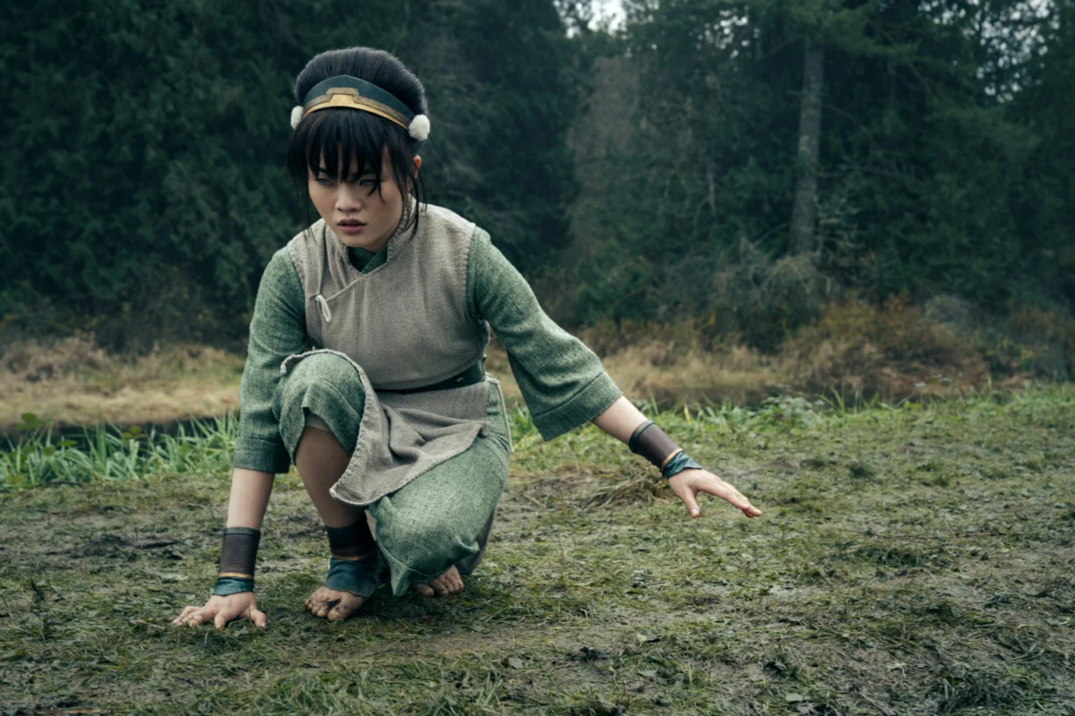 "A young girl with black hair wearing green clothes crouches down in the woods with bare feet on grass and right hand touching the ground. "