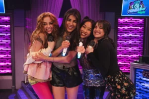 "Four young women smiling and hugging while holding microphones, standing together on a colorful karaoke stage decorated with purple lights and a "Tunes-day Night" sign, suggesting a lively and fun indoor environment."