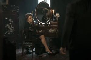 "Woman in dark robe sits at vintage vanity with mirror and candles in dimly lit, elegant room; another person stands in foreground, partly visible."
