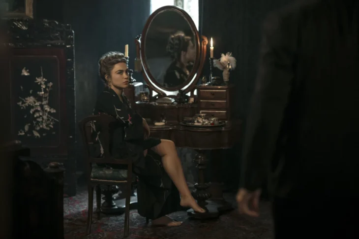 "Woman in dark robe sits at vintage vanity with mirror and candles in dimly lit, elegant room; another person stands in foreground, partly visible."