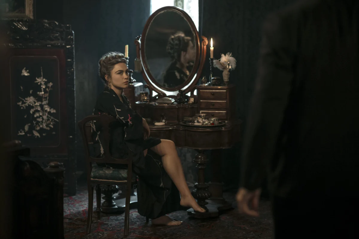 "Woman in dark robe sits at vintage vanity with mirror and candles in dimly lit, elegant room; another person stands in foreground, partly visible."