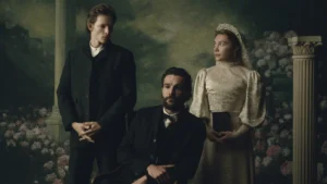 "Three people in Victorian-era clothing pose in a formal portrait setting with floral and classical column background; one man seated, one man standing, and a woman in a white dress holding a book."