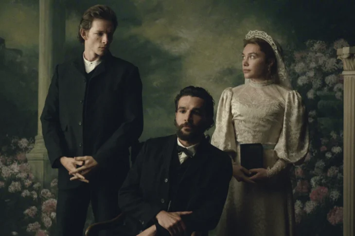 "Three people in Victorian-era clothing pose in a formal portrait setting with floral and classical column background; one man seated, one man standing, and a woman in a white dress holding a book."