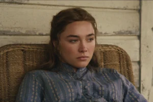 "A young woman in a blue patterned dress sits pensively in a wicker chair against a worn wooden wall, appearing thoughtful and somber in a rustic, vintage setting."