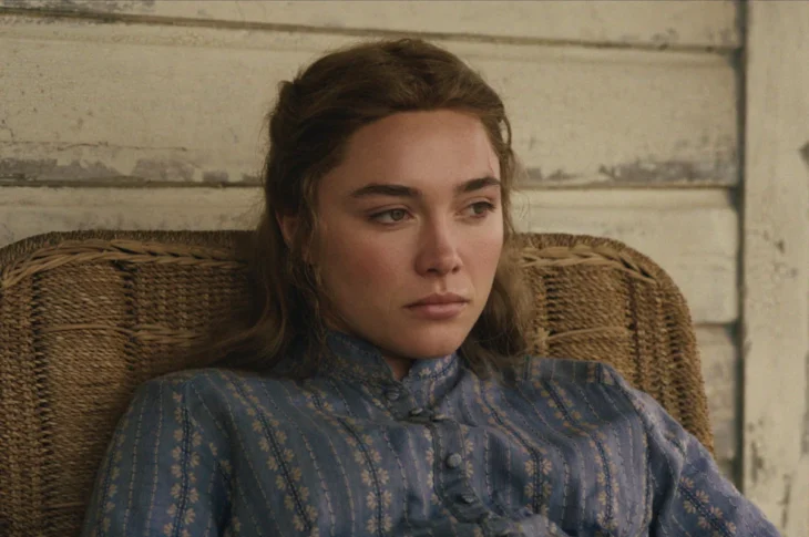 "A young woman in a blue patterned dress sits pensively in a wicker chair against a worn wooden wall, appearing thoughtful and somber in a rustic, vintage setting."