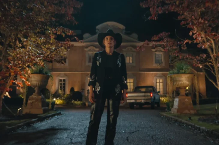 "Person in a black cowboy outfit stands on a driveway at night, with autumn trees on both sides and a large, well-lit house and parked cars in the background."