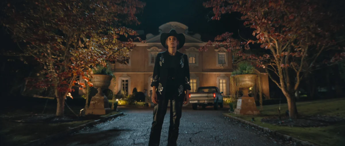 "Person in a black cowboy outfit stands on a driveway at night, with autumn trees on both sides and a large, well-lit house and parked cars in the background."
